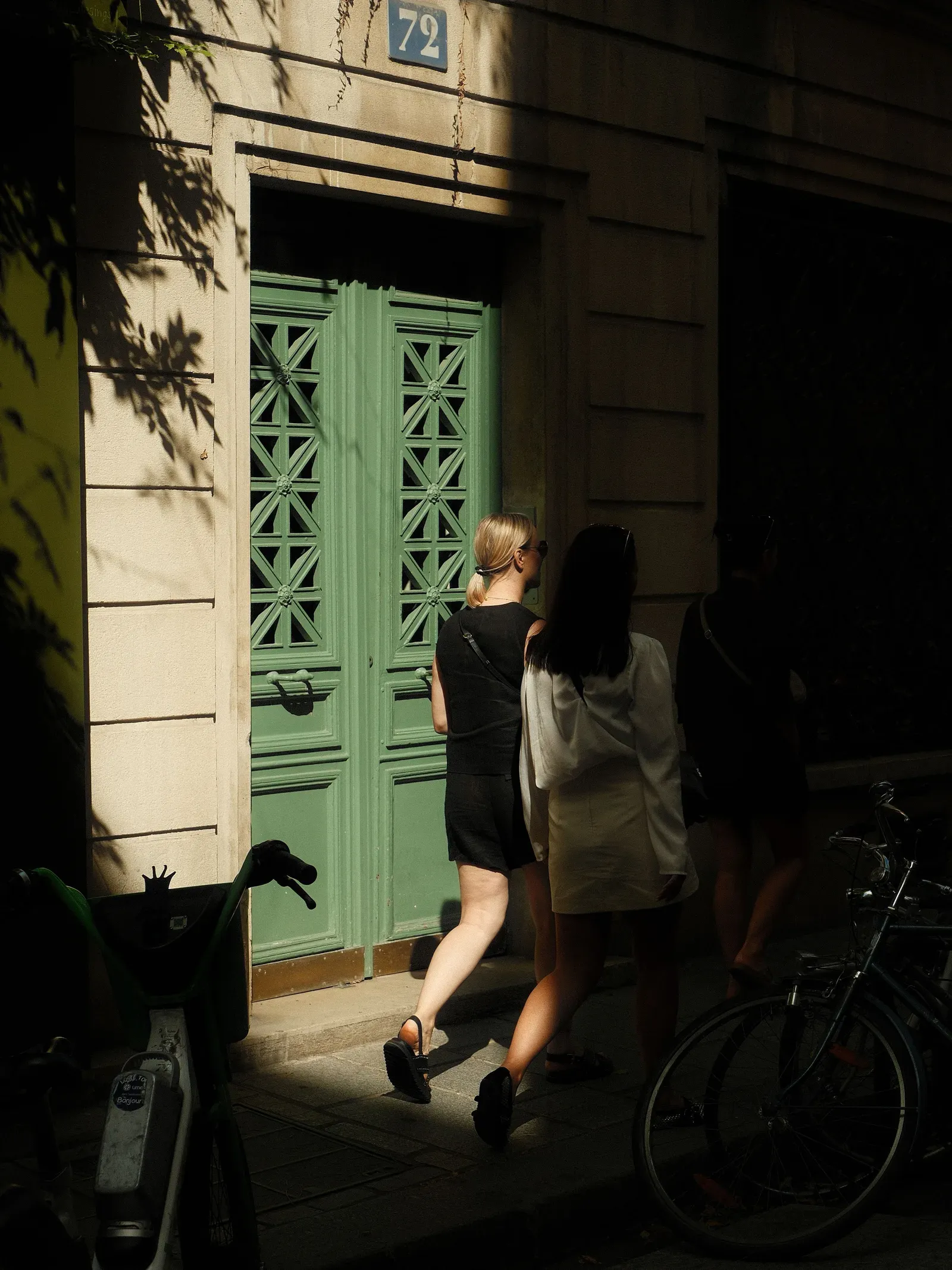 Deux personnes passent devant une grande porte en bois vert aux panneaux ajourés, dans un immeuble en pierre baigné de soleil. Des vélos sont garés à l’ombre.
